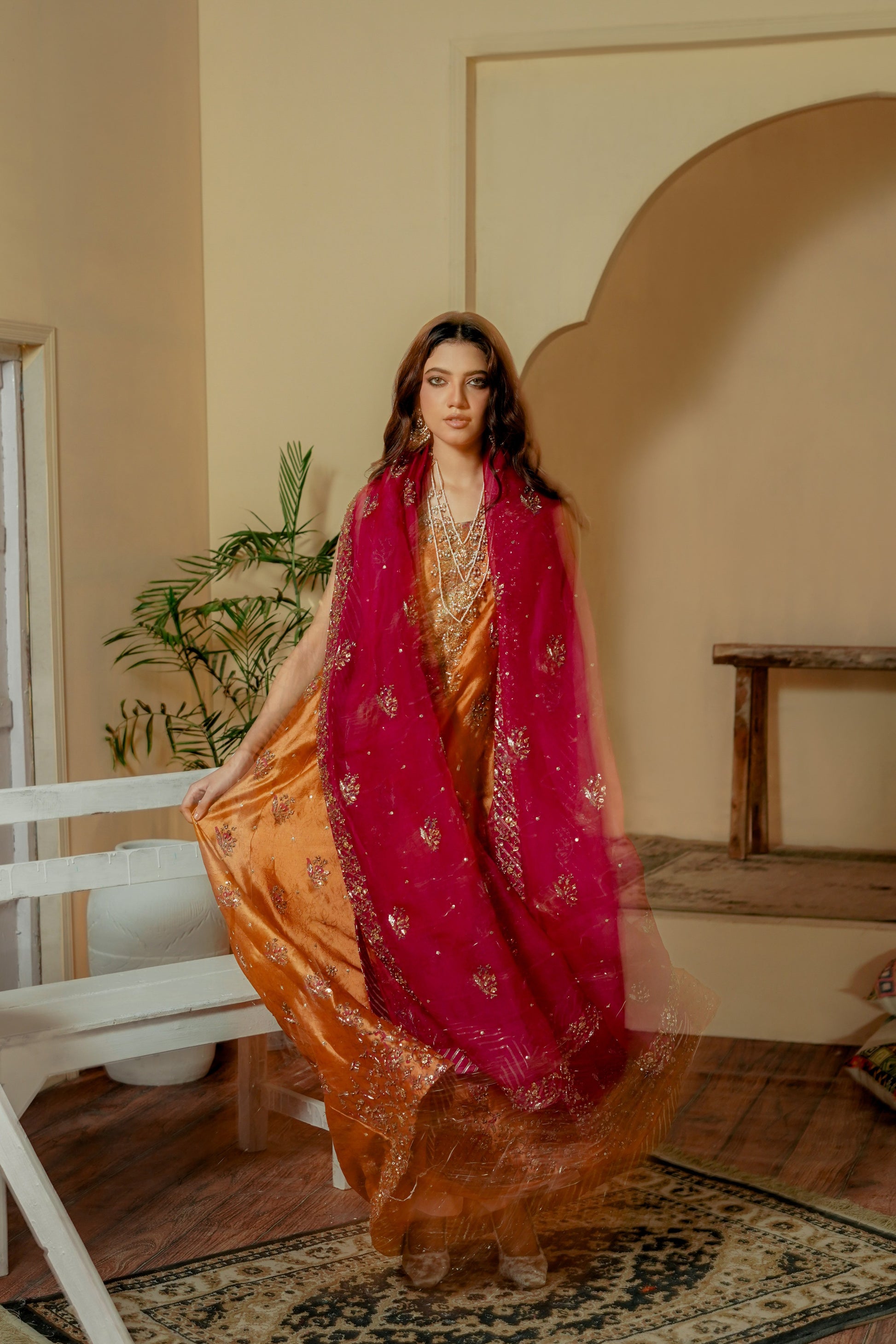 Woman in a traditional formal wear maxi outfit with a red dupatta in a room with a plant and wooden table.