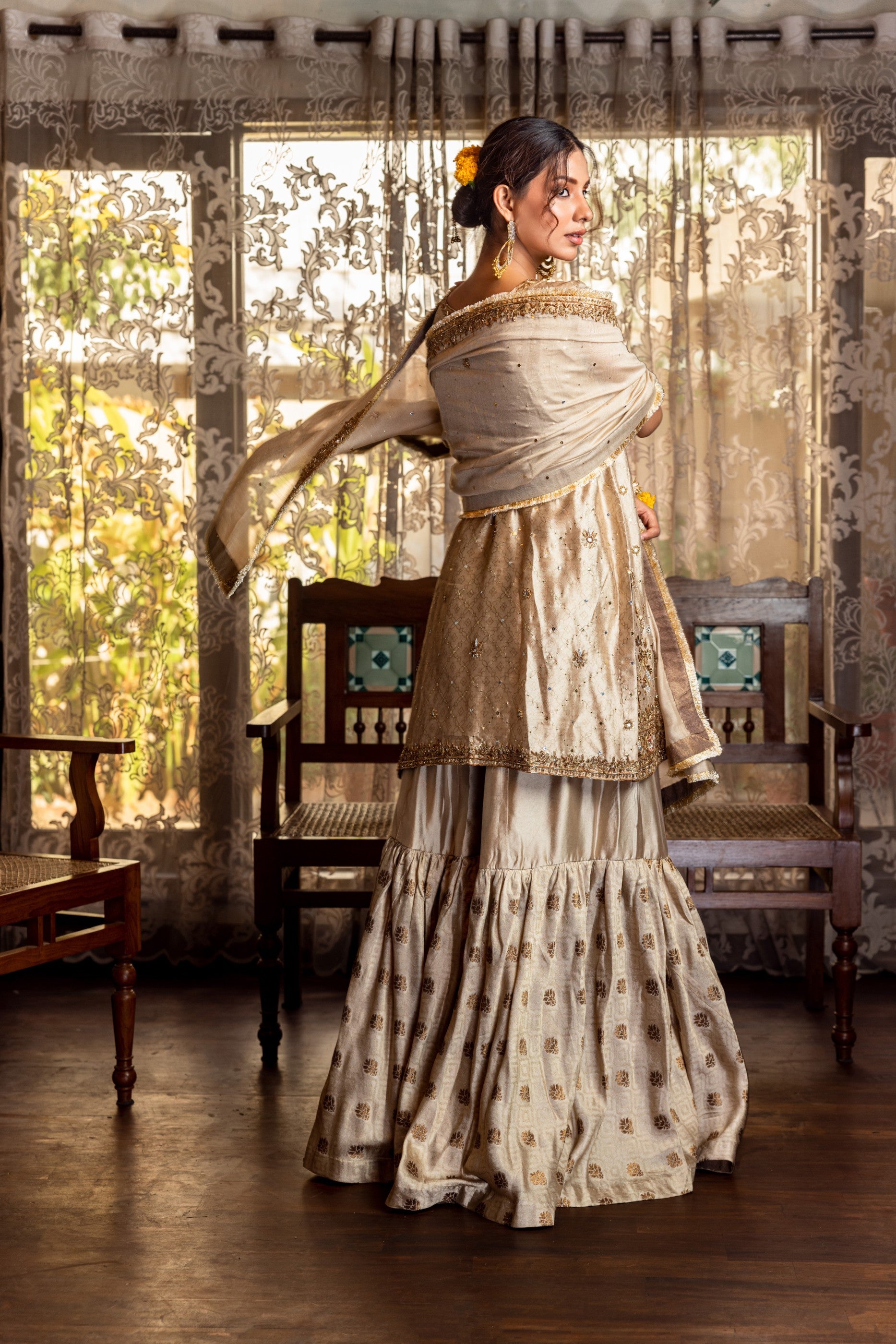 Woman in traditional Party Wear Garara  standing in a room with wooden furniture and curtains.