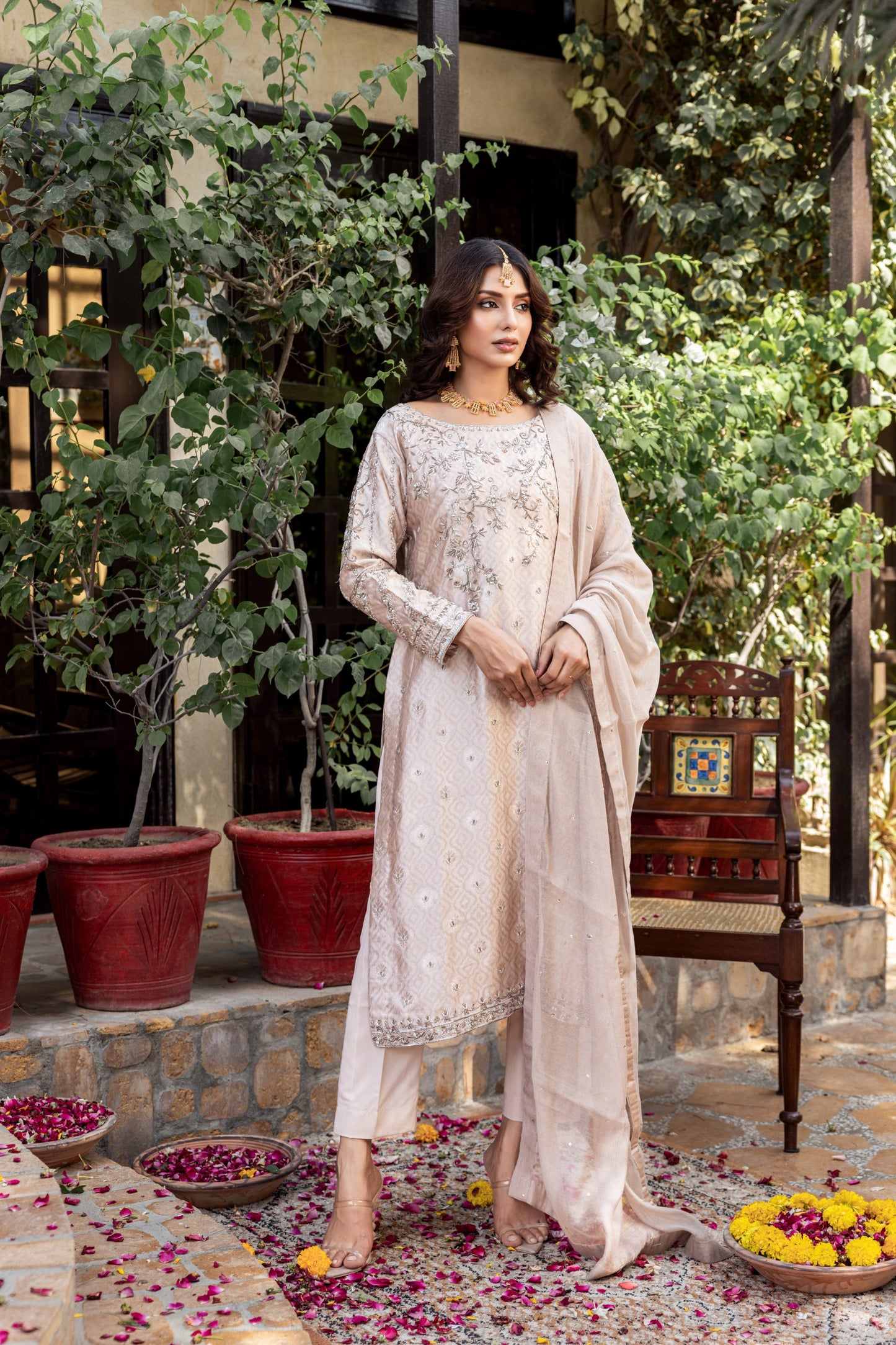 Woman in a beige embroidered formal basic shirt outfit standing in a garden setting with potted plants and a chair.