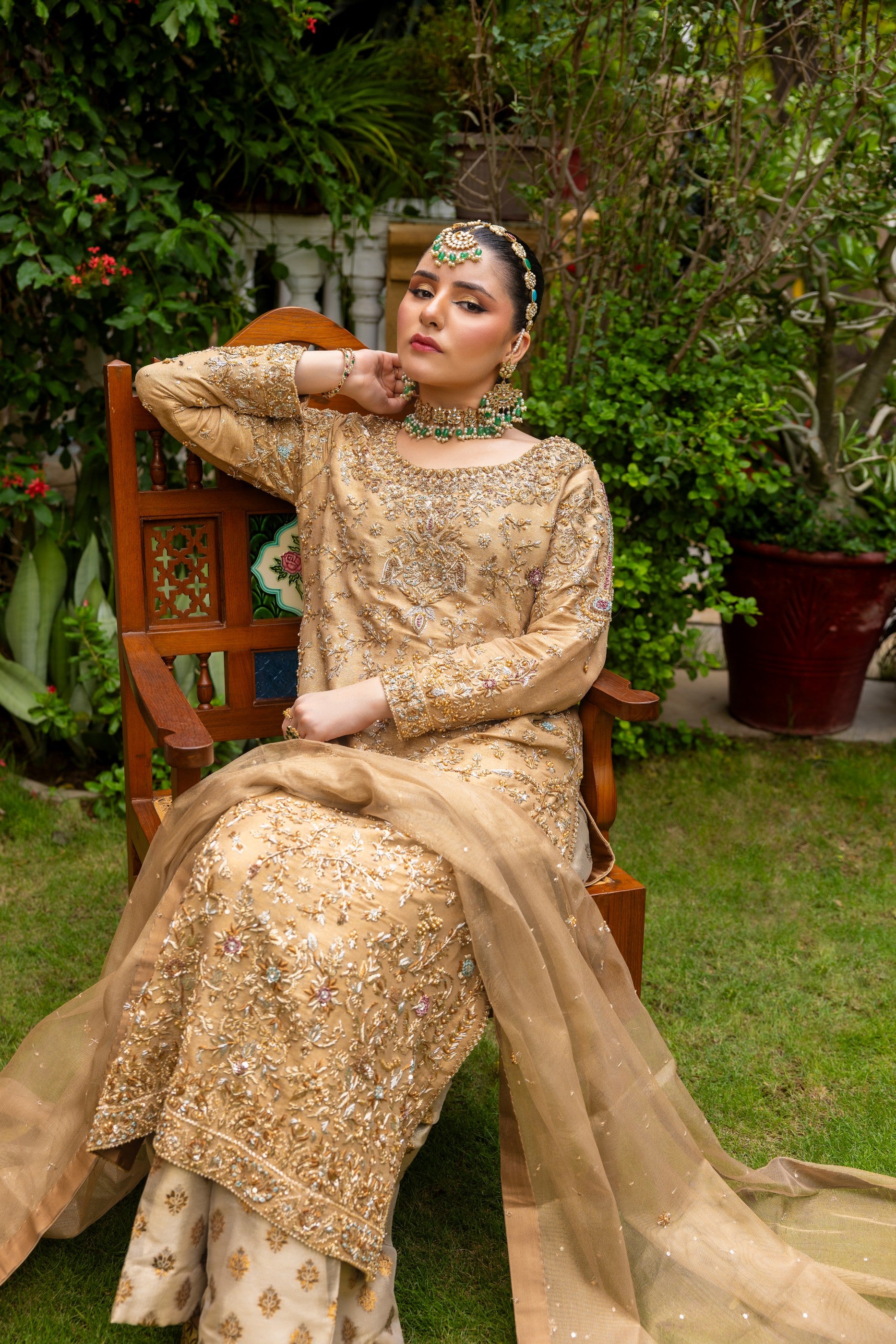 Woman in a gold embroidered traditional party wear shirt outfit sitting outdoors with greenery in the background