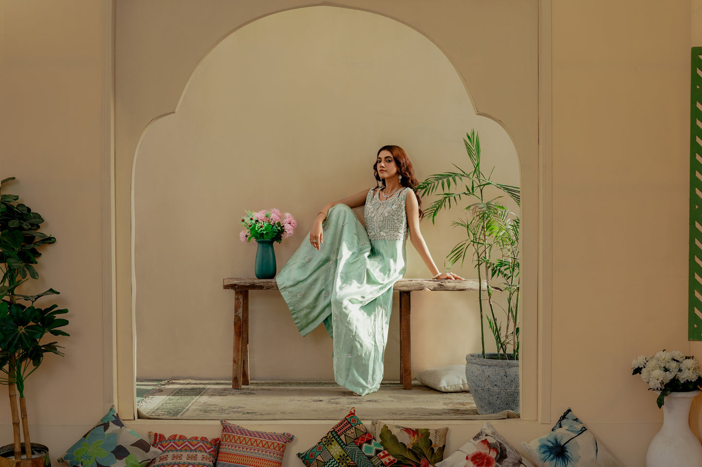 Woman in a light green party wear maxi sitting on a wooden bench in a decorative room with plants and a vase.