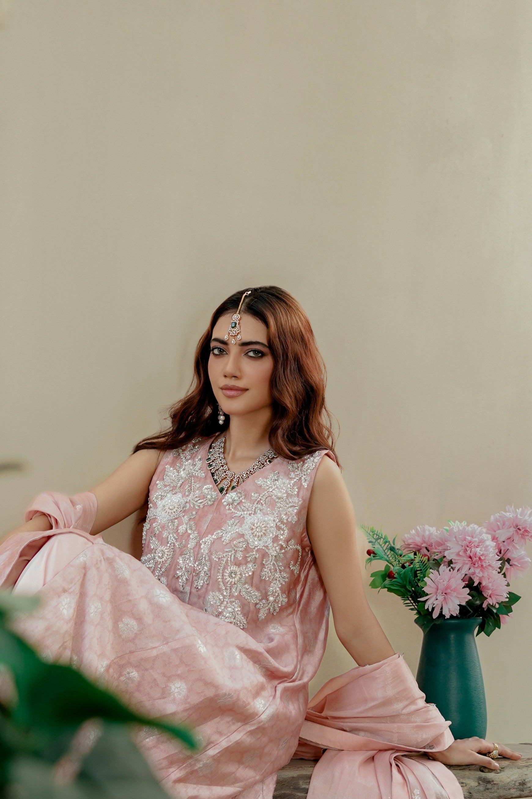 Woman in a pink traditional formal maxi outfit with floral patterns, sitting indoors with a neutral background.