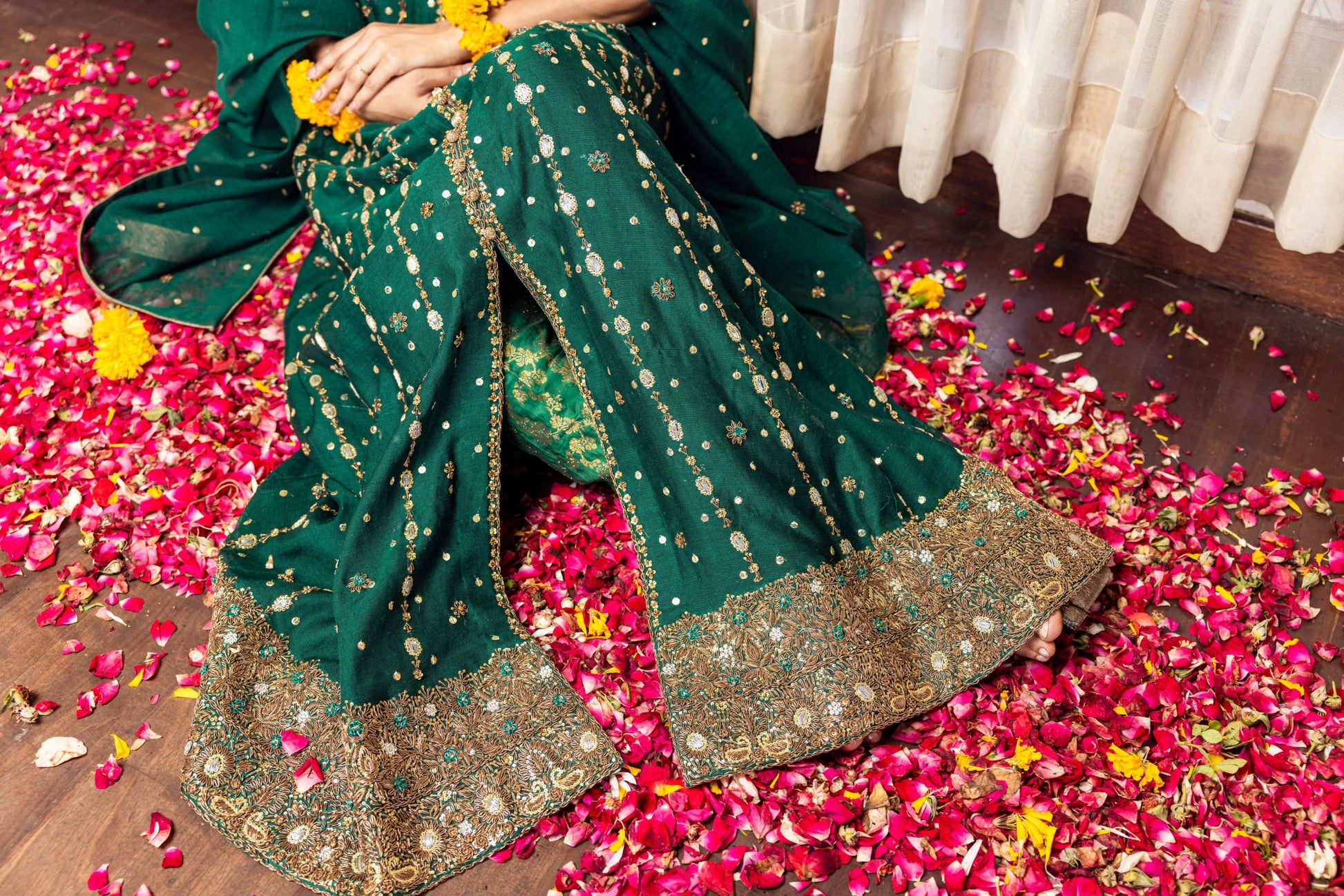 Person wearing a green traditional front-open maxi outfit with gold embroidery on a floor scattered with rose petals.