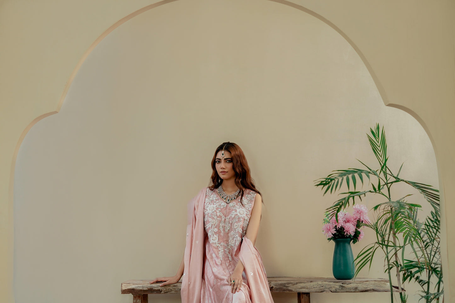 Woman in a pink traditional formal maxi outfit standing in front of a decorative archway with a vase of flowers and plants.