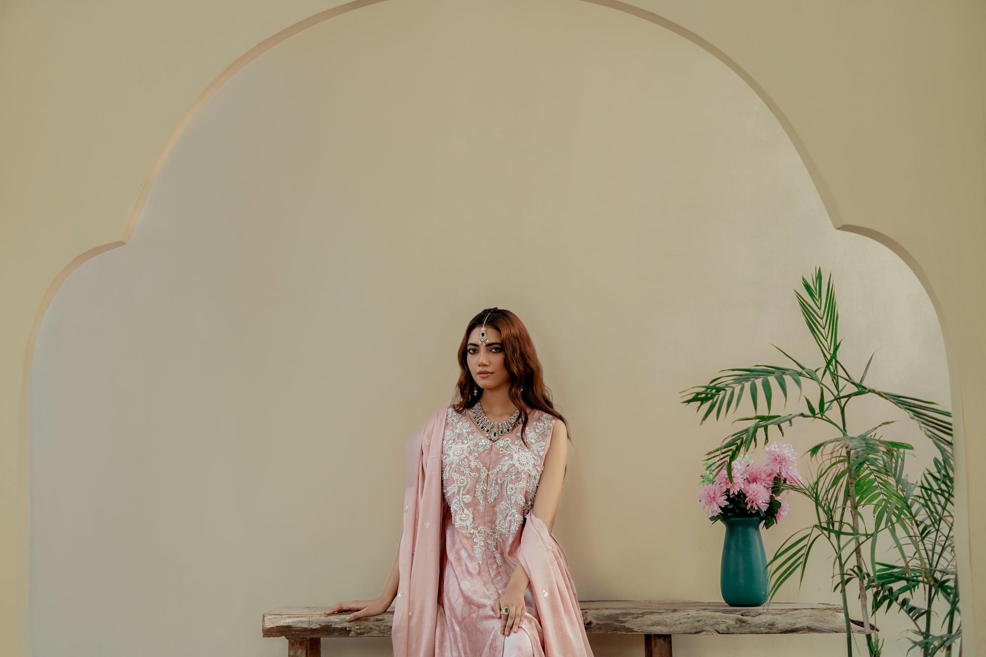 Woman in a pink traditional formal maxi outfit standing in front of a decorative archway with a vase of flowers and plants.