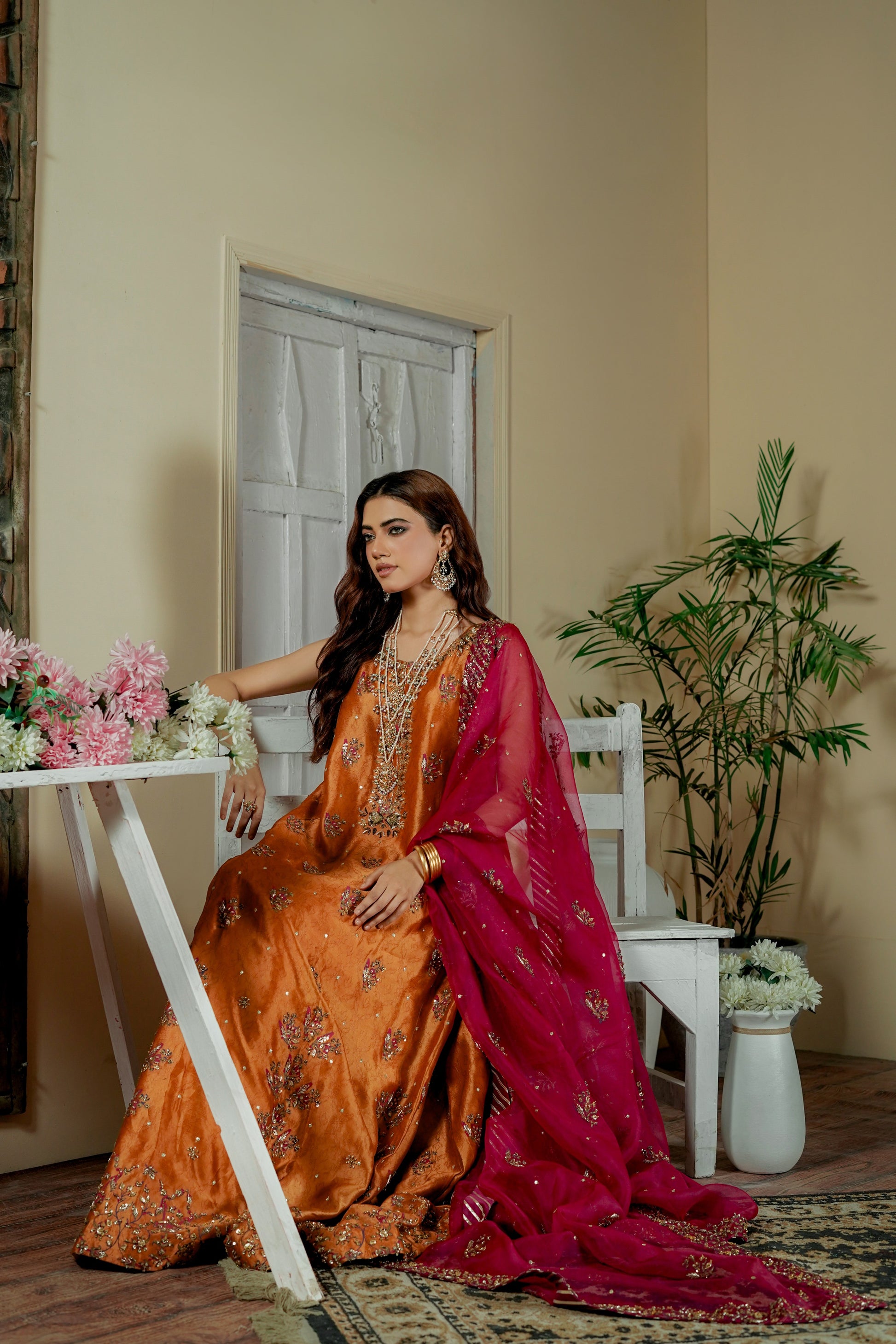 Woman in traditional orange and red formal wear maxi outfit sitting in a room with decor elements.
