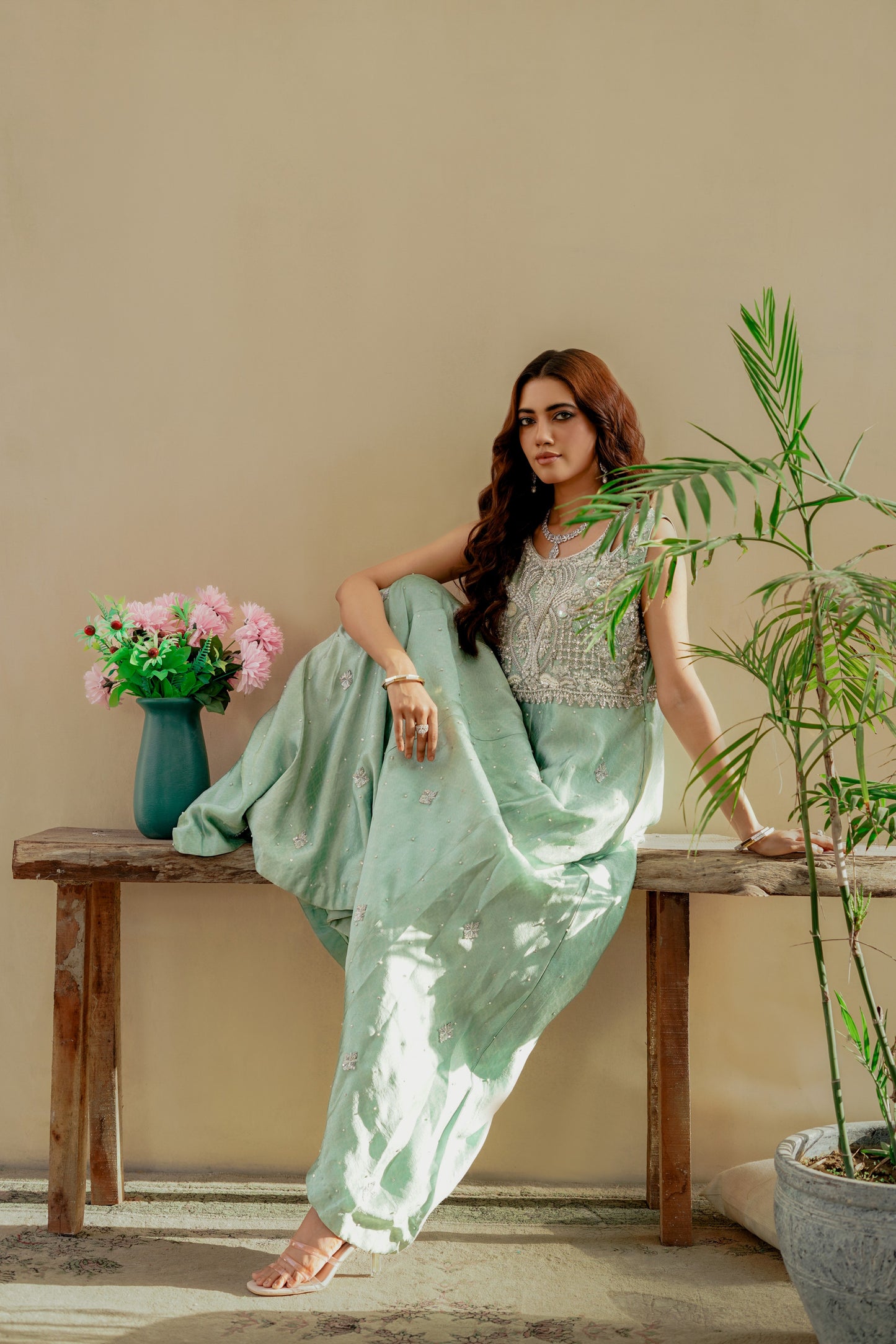 Woman in a light green party wear maxi outfit sitting on a wooden bench with plants and flowers in the background.