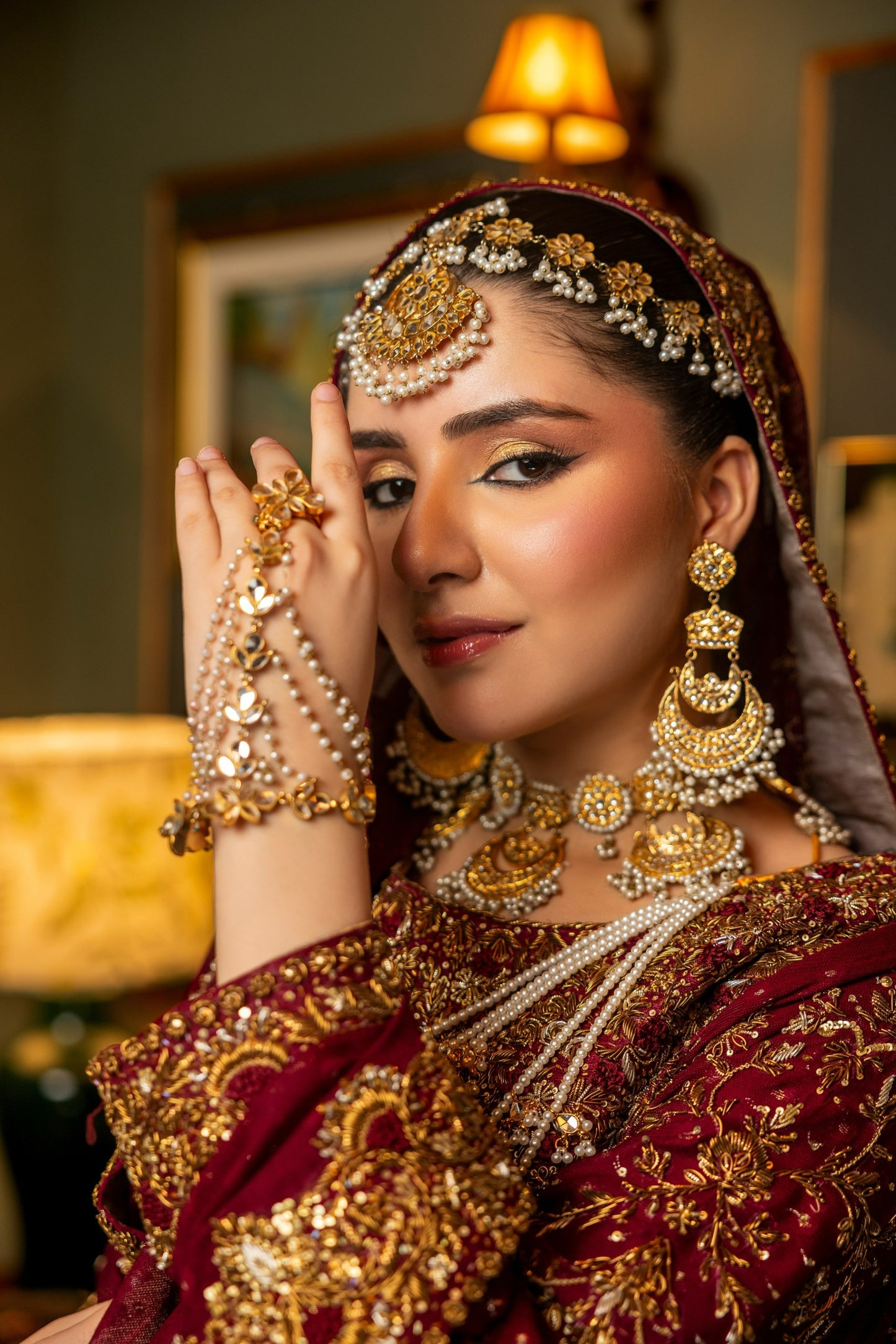 Woman in traditional bridal sharara attire with gold jewelry and a blurred indoor background