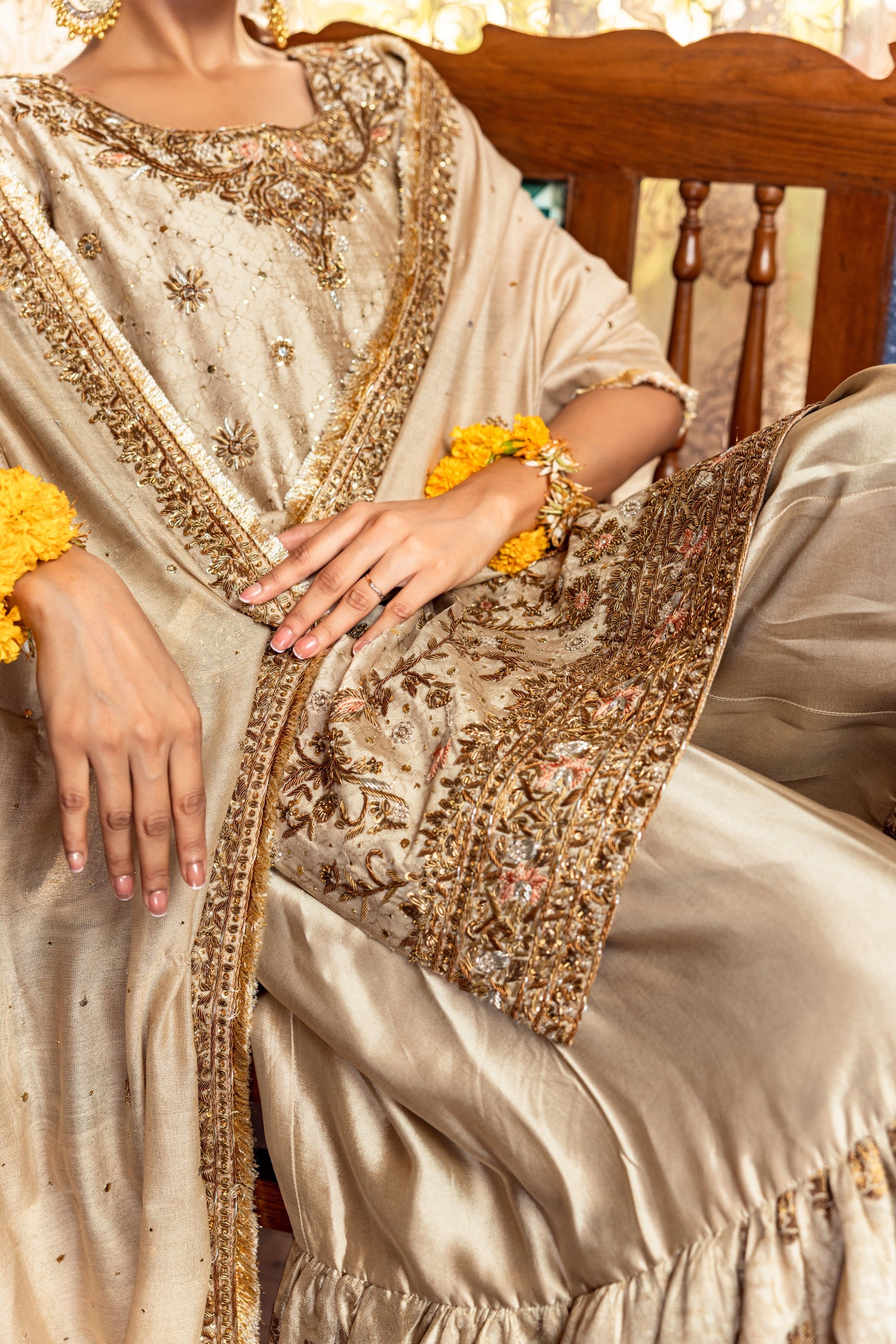 Person wearing a cream-colored Party Wear Garara with gold embroidery, sitting on a wooden chair.