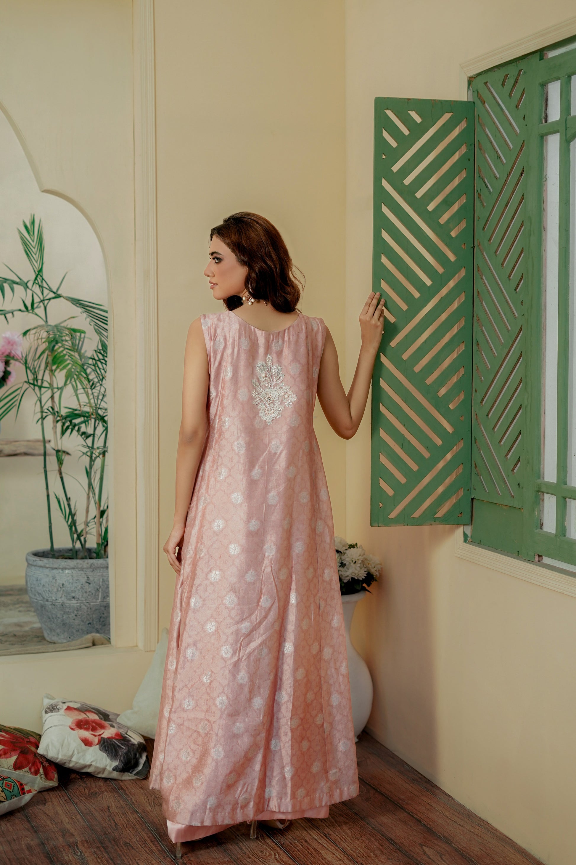 Woman in a pink formal maxi dress standing in a room with green shutters and plants.