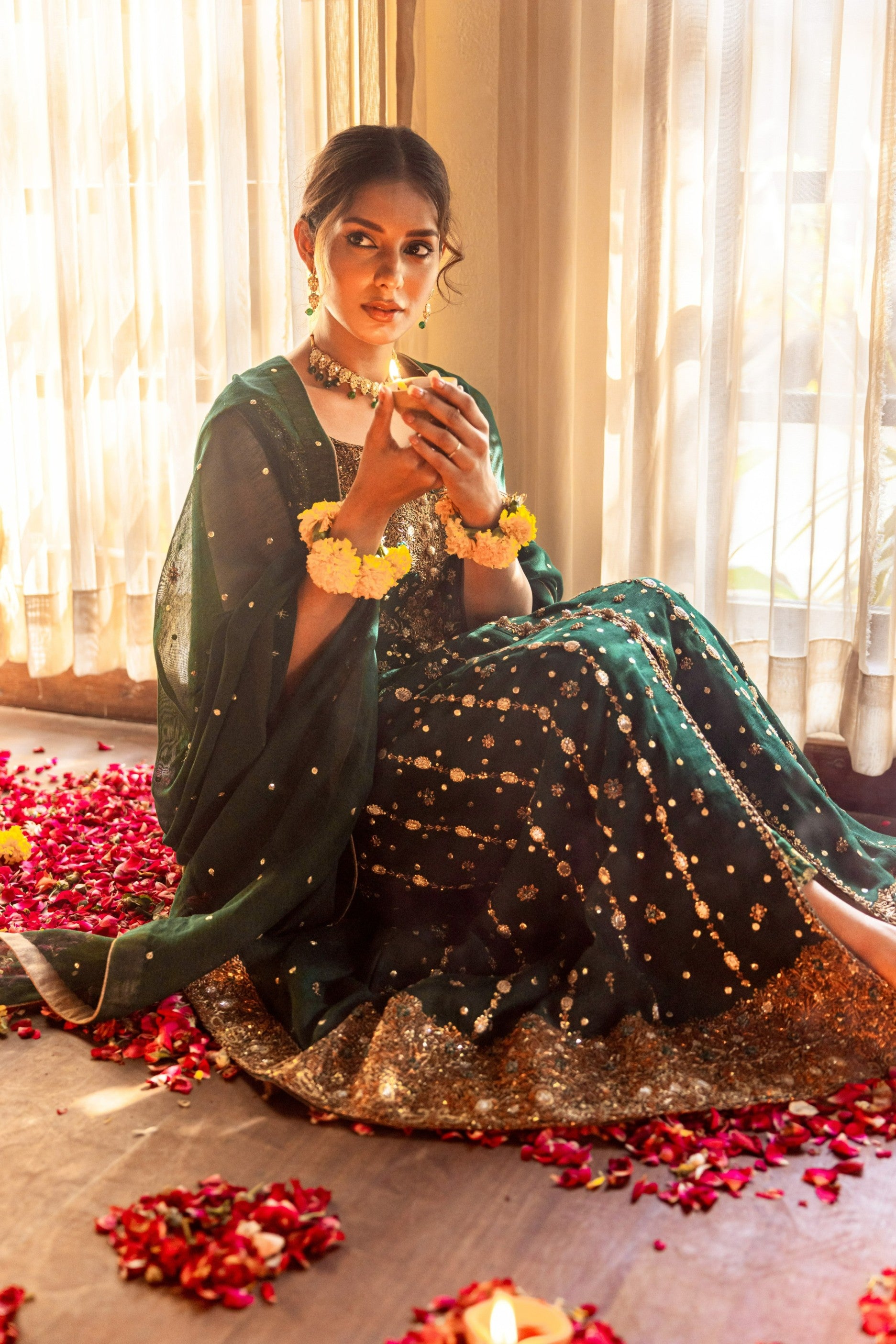 Woman in traditional green and gold front-open maxi outfit sitting on the floor with floral decorations.