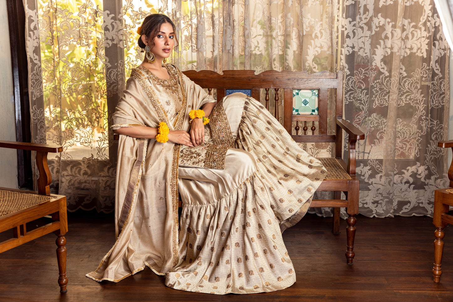 Woman in a traditional Party Wear Garara sitting on a wooden chair with floral curtains in the background