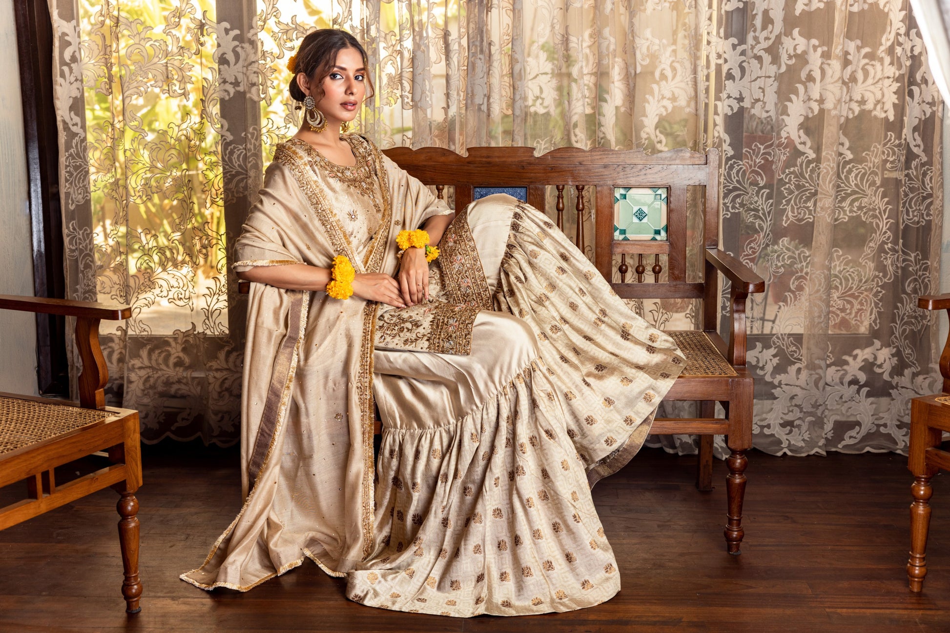 Woman in a traditional Party Wear Garara sitting on a wooden chair with floral curtains in the background