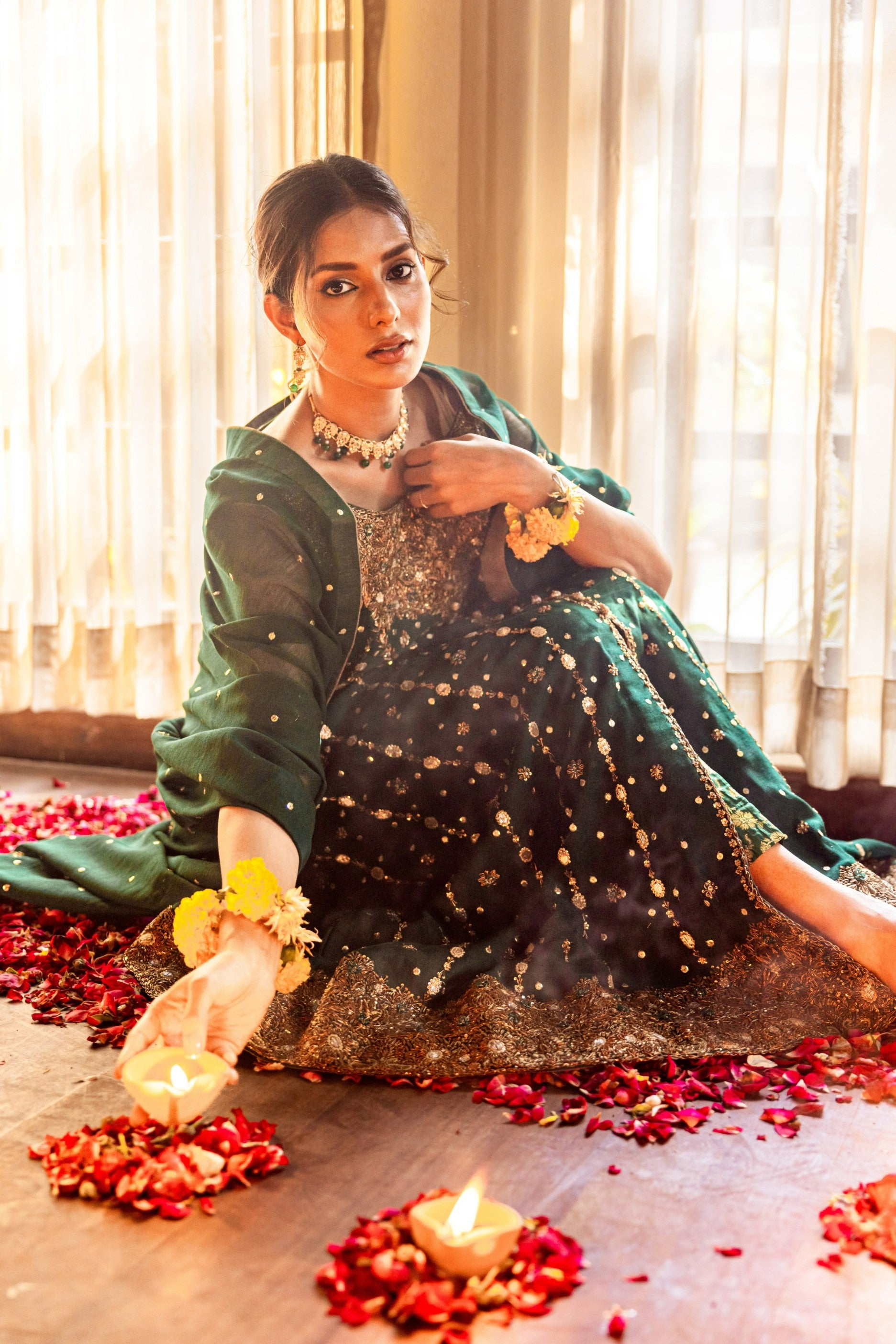 Woman in traditional front-open maxi sitting on a floor with candles and flowers, surrounded by warm lighting.