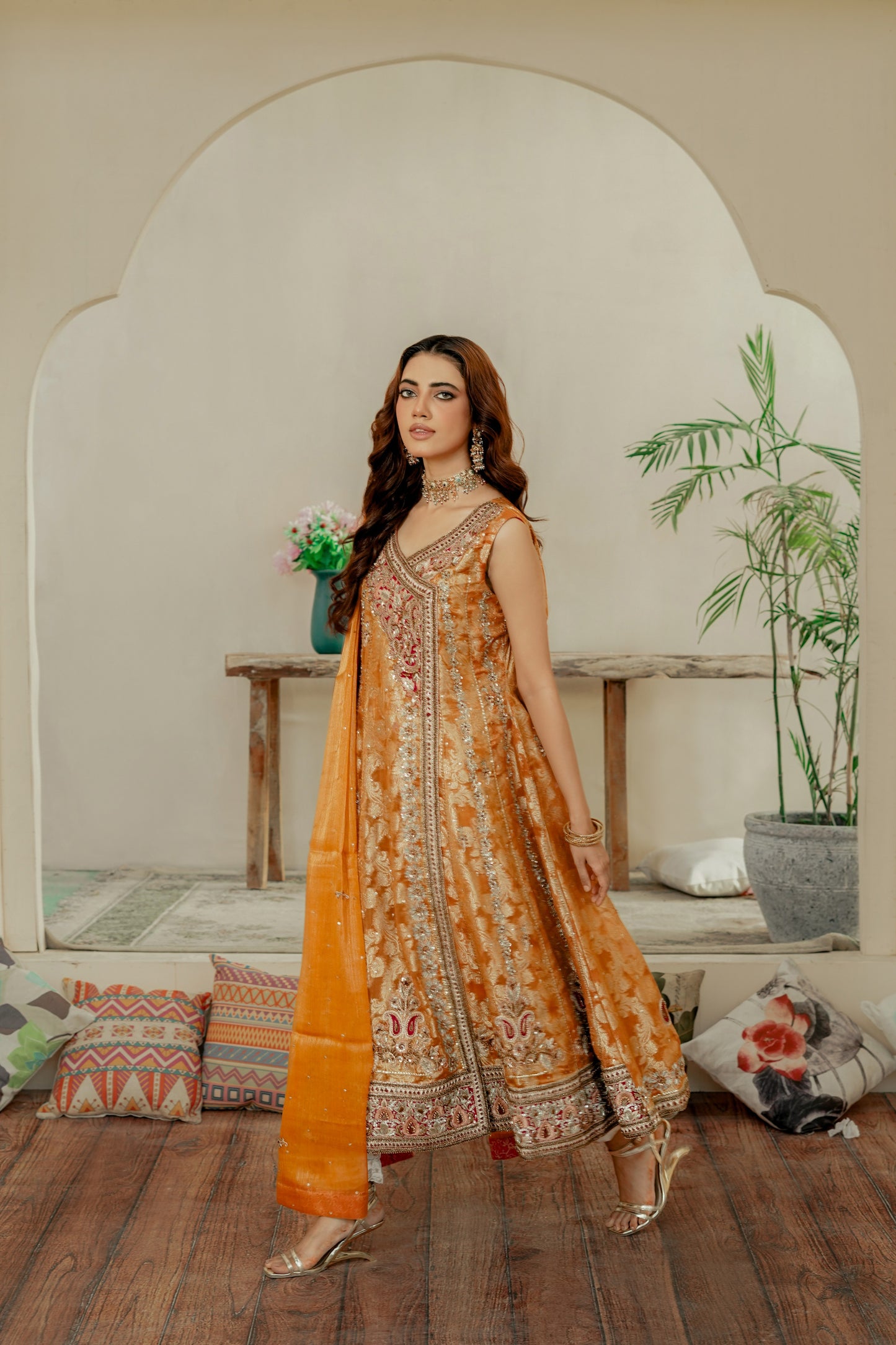 Woman in a traditional formal frock outfit standing in a room with wooden floor and decorative pillows.
