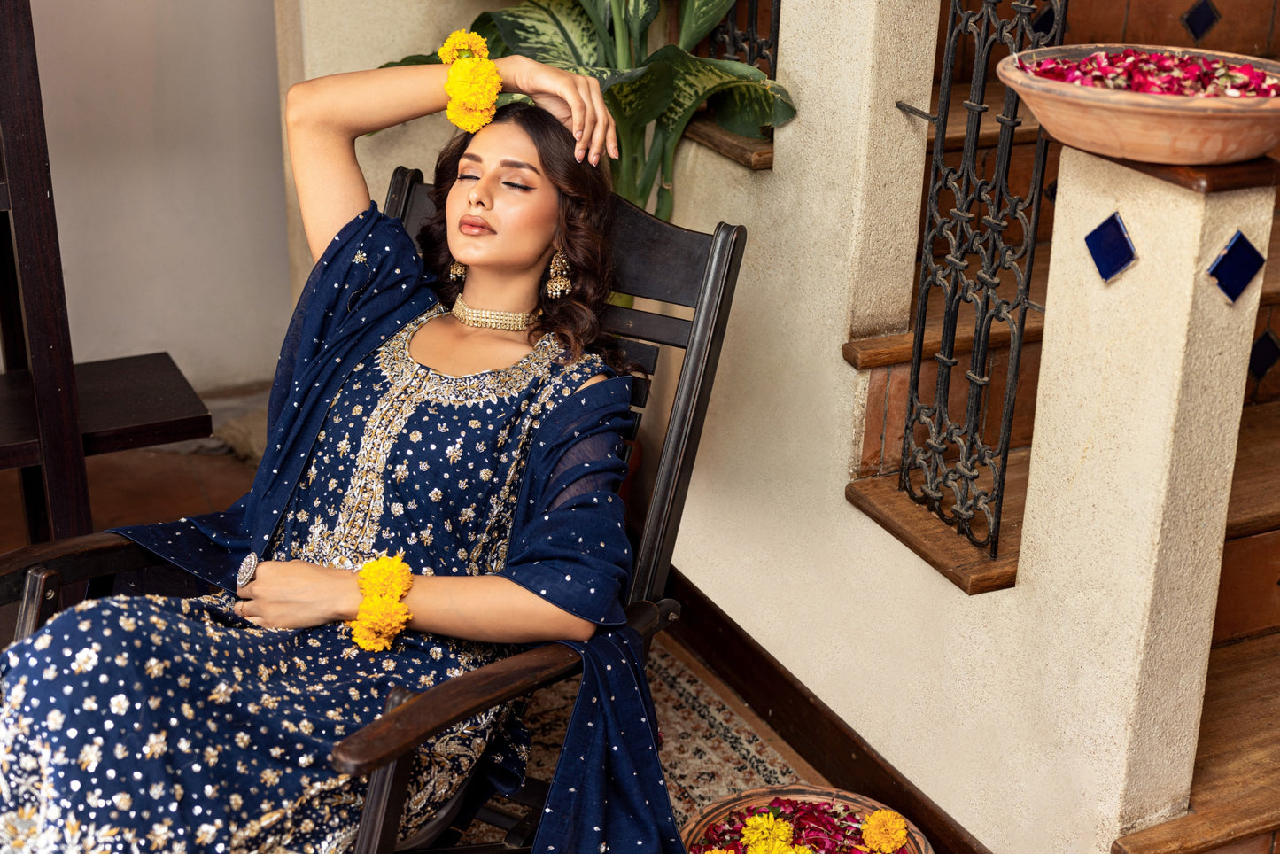 Woman in a blue traditional formal outfit sitting on a chair with flowers in her hair and hands.