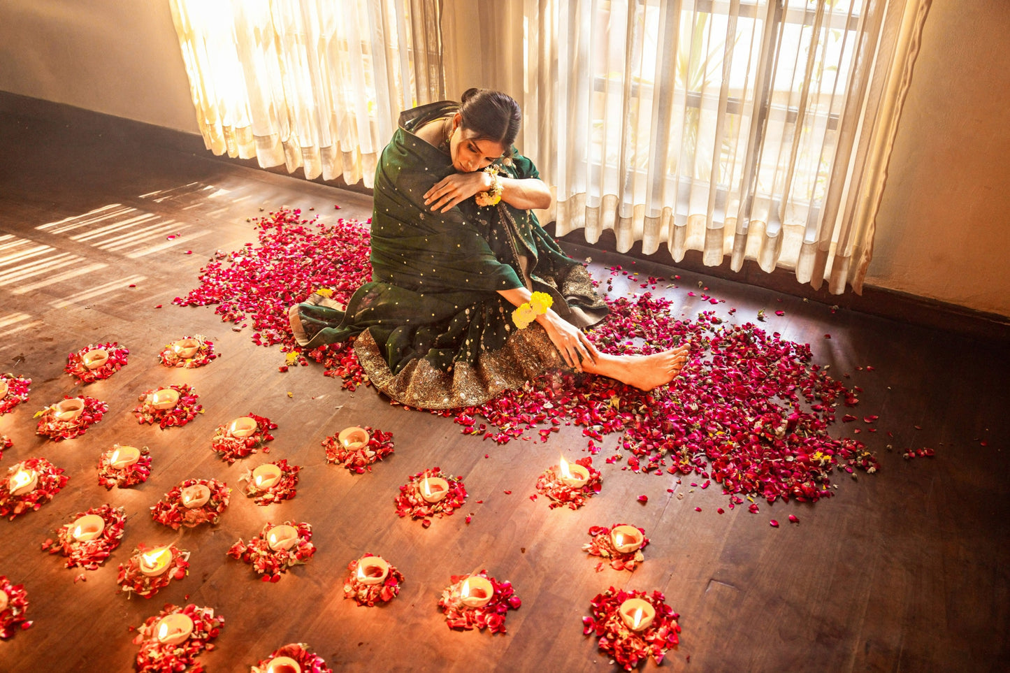 Woman sitting on the floor surrounded by red flowers and candles, with curtains in the background.