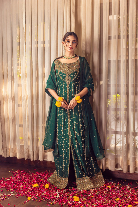 Woman in a green traditional front-open maxi outfit standing in front of curtains with flower petals on the floor.