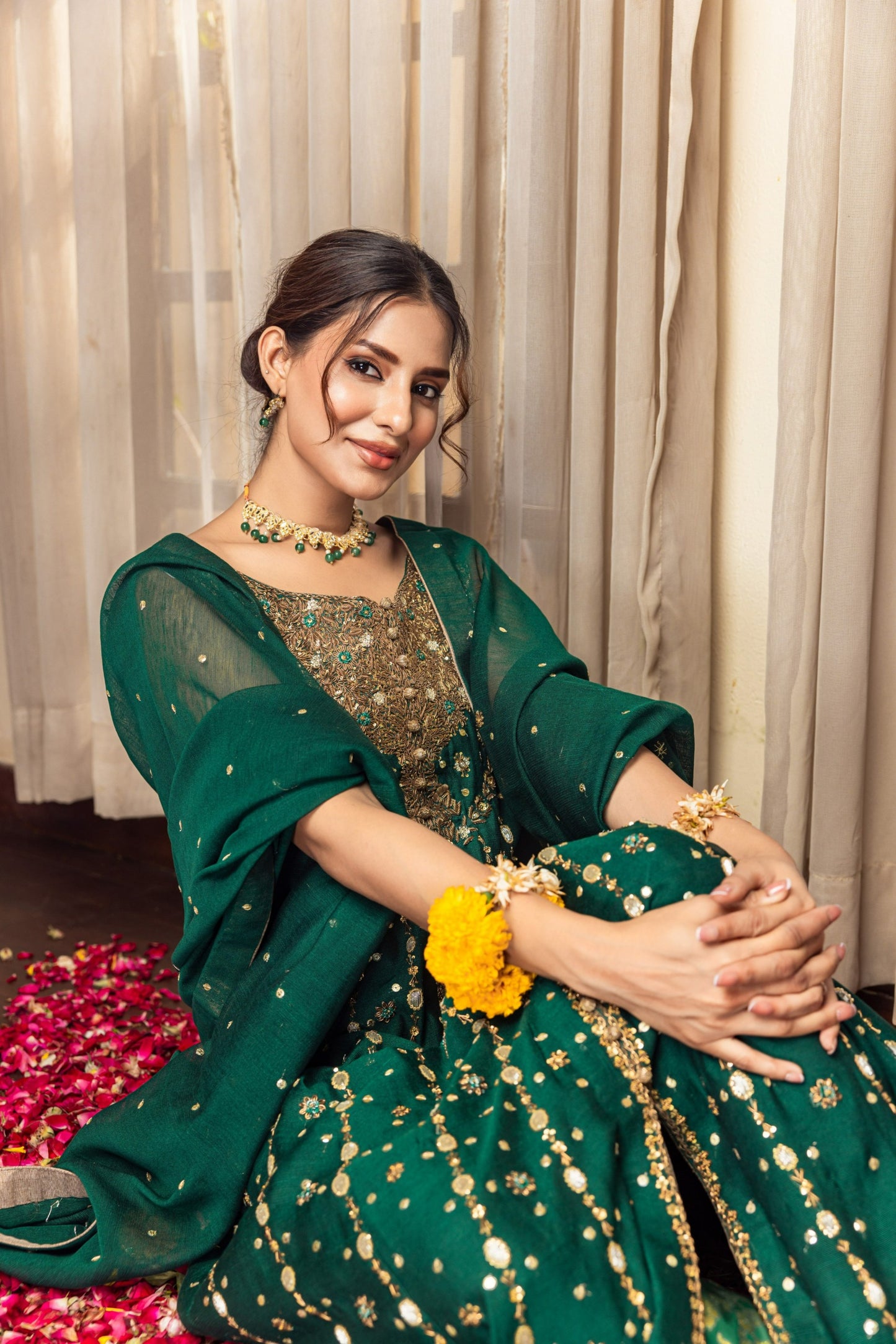 Woman in a green traditional front-open maxi outfit with gold details sitting against a neutral curtain background.