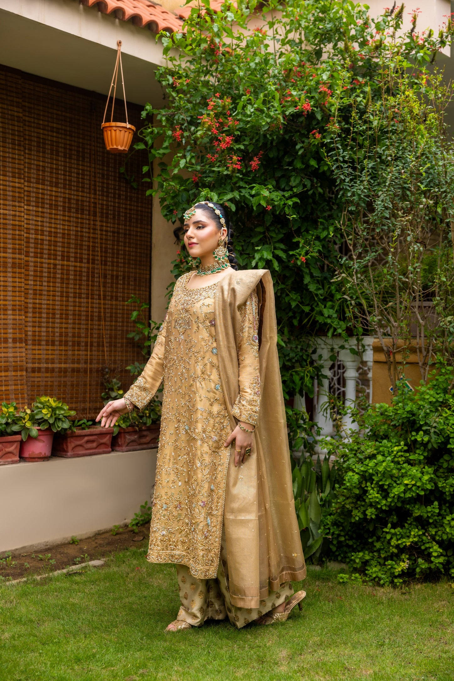 Woman in a beige traditional party wear shirt standing outdoors with greenery and a house in the background