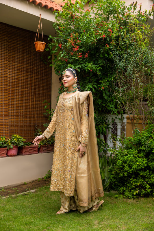 Woman in a beige traditional party wear shirt standing outdoors with greenery and a house in the background