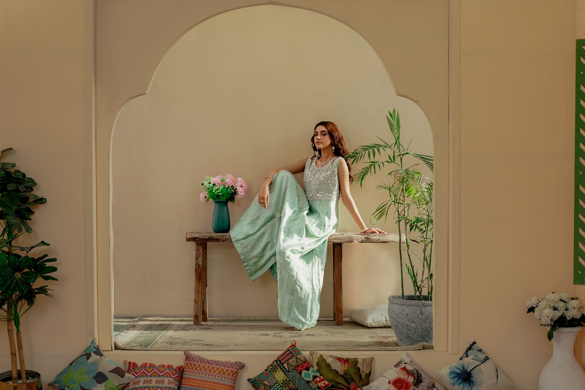 Woman in a light green party wear maxi sitting on a wooden bench in a decorative room with plants and a vase.