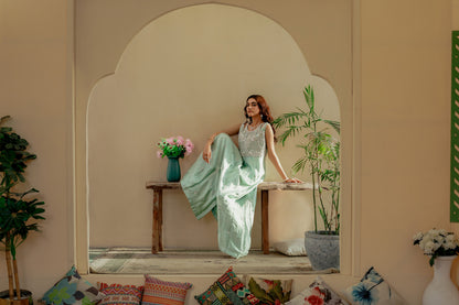 Woman in a light green party wear maxi sitting on a wooden bench in a decorative room with plants and a vase.