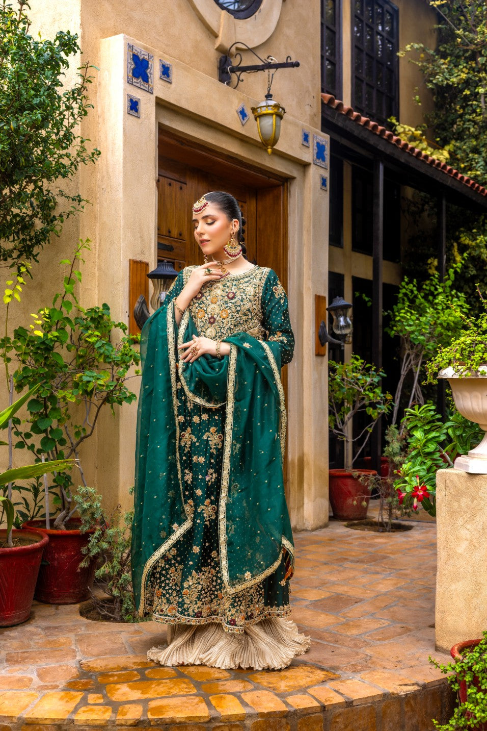 Woman in a green front-open maxi traditional outfit standing outdoors with plants and a building in the background.