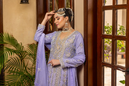 Woman in a light purple bridal maxi outfit standing indoors with plants and a door in the background.