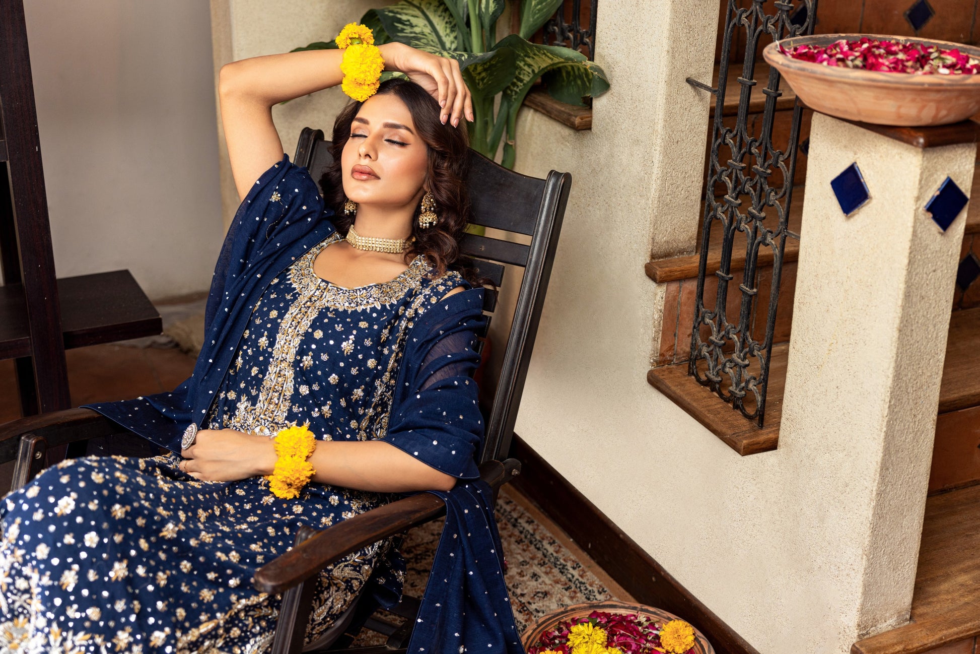 Woman in a blue traditional formal outfit sitting on a chair with flowers in her hair and hands.