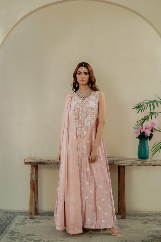 Woman in a pink floral formal maxi dress standing in a room with a wooden bench and vase of flowers.