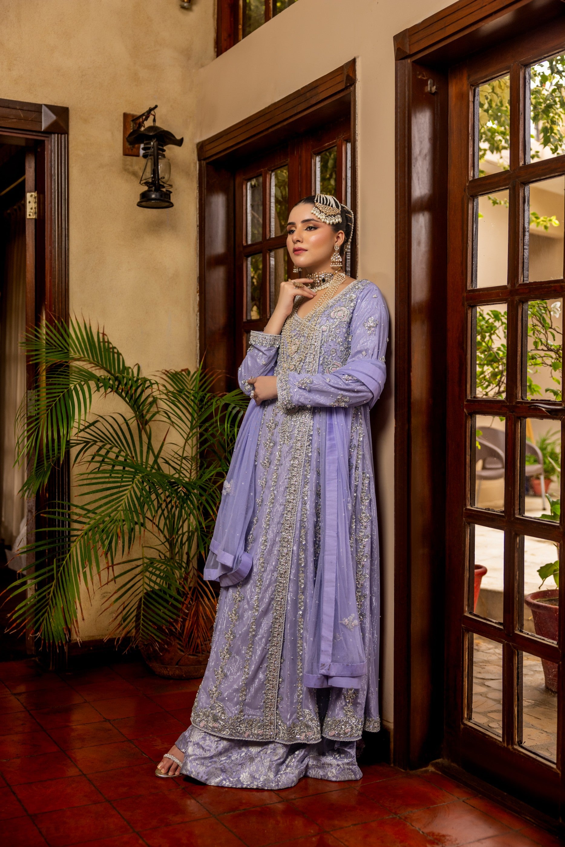 Woman in a light purple traditional bridal maxi outfit standing in a doorway with plants and a door in the background.