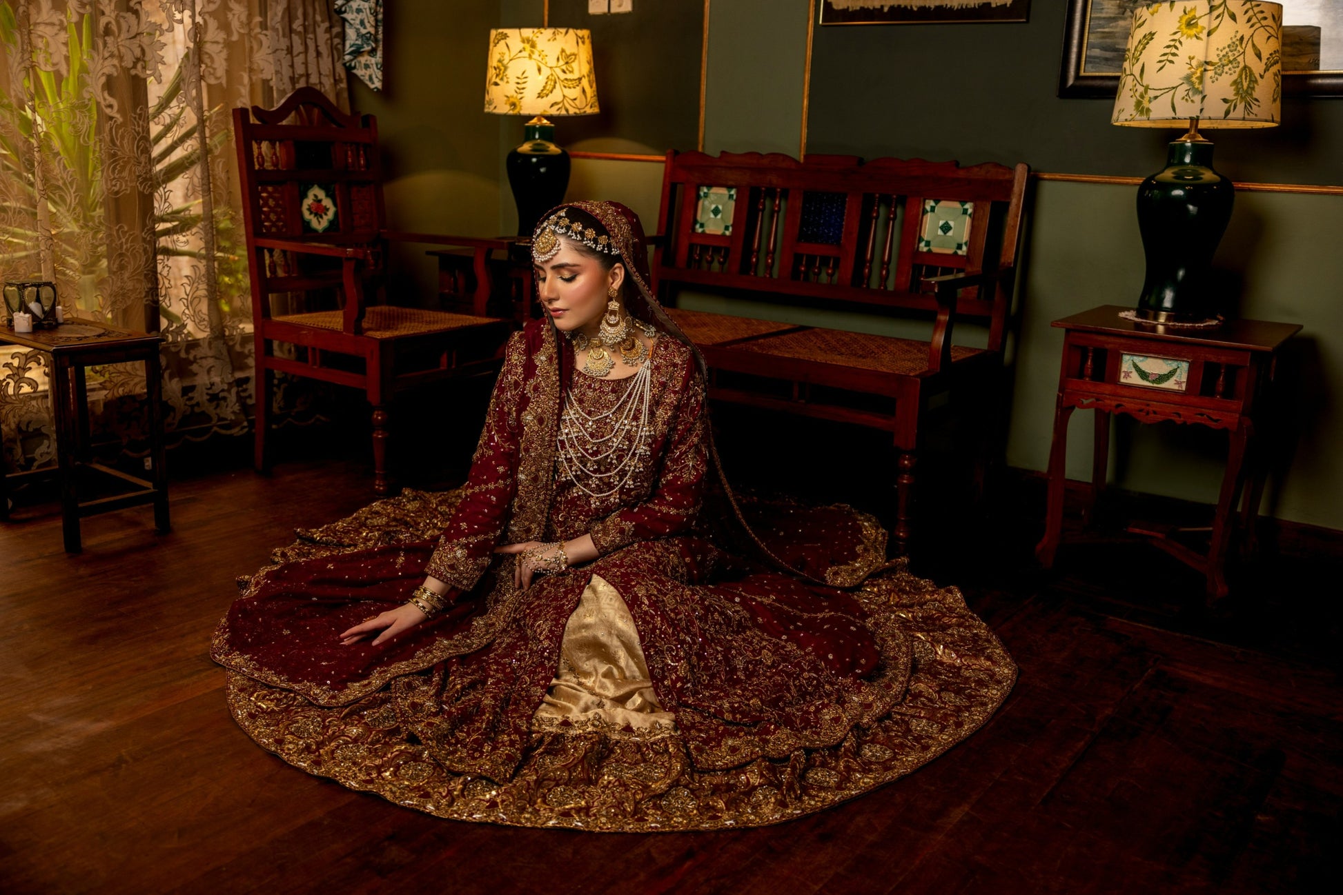 Woman in traditional bridal sharara attire sitting on a rug in a dimly lit room with wooden furniture and lamps.