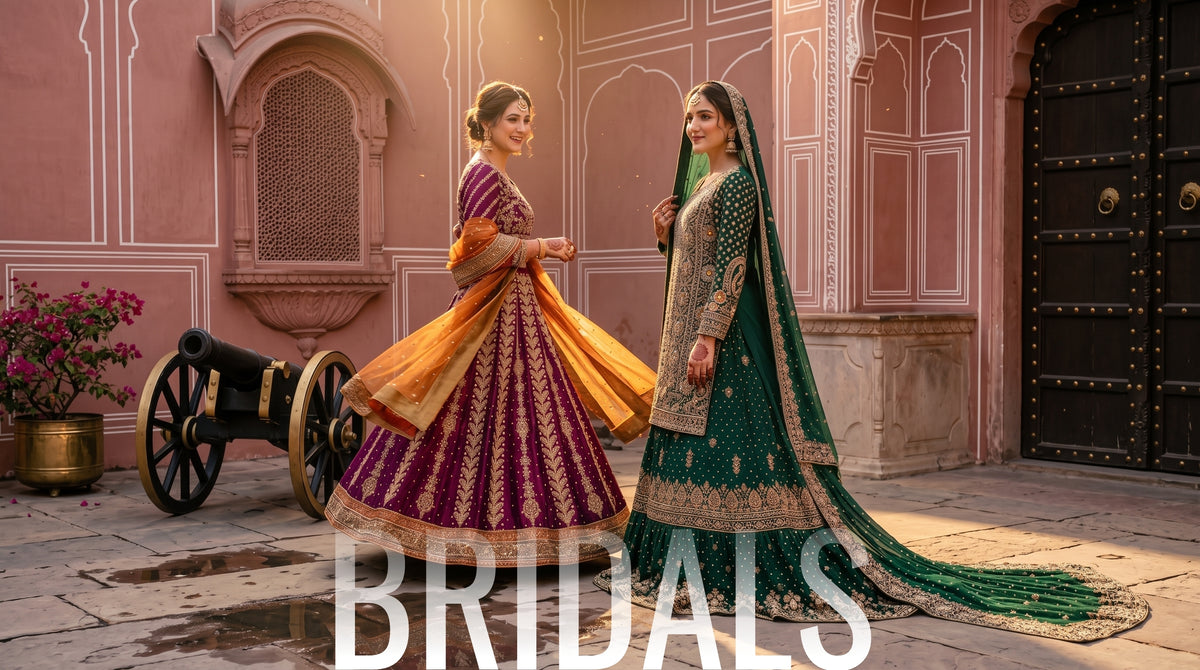 Two women in traditional Indian bridal attire standing in front of a pink palace wall. One wears a purple and orange lehenga with gold embroidery, and the other wears a green and gold lehenga with a long dupatta.